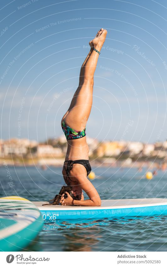 Slim woman in swimwear doing headstand on paddleboard sup board yoga sea water tranquil asana ripple aqua female slim practice balance pose position mindfulness