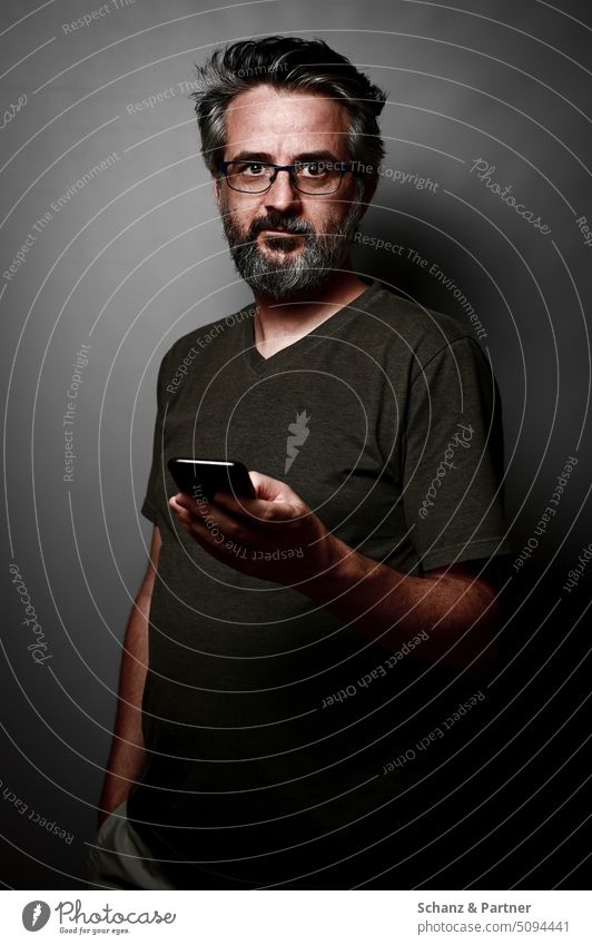 bearded man with glasses in t-shirt with smartphone against gray monochrome background looks at camera Earnest Dark Shadow masculine Rich in contrast Eyeglasses