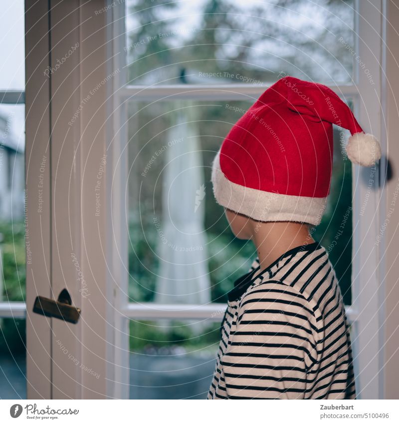 Little boy with red Christmas or Santa Claus cap in striped pajamas stands at window waiting for Christmas Boy (child) Small Cap Wait Red White Window look out