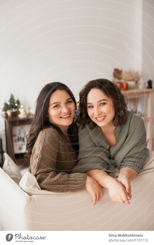 Two women sitting on a couch at cozy home apartment. Family spending time at home. Portrait of sisters smiling in modern minimal interior. comfortable