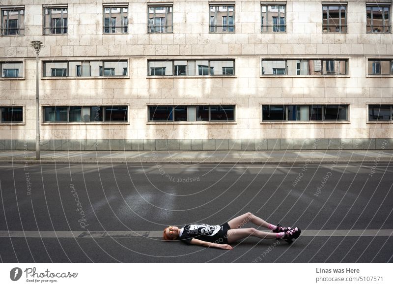 A fashionable street in Berlin where a red hair model is lying on the floor while being all gorgeous. Concrete surrounds her. Her high heels, pink socks, black clothes, and fancy sunglasses accompany her killer look.