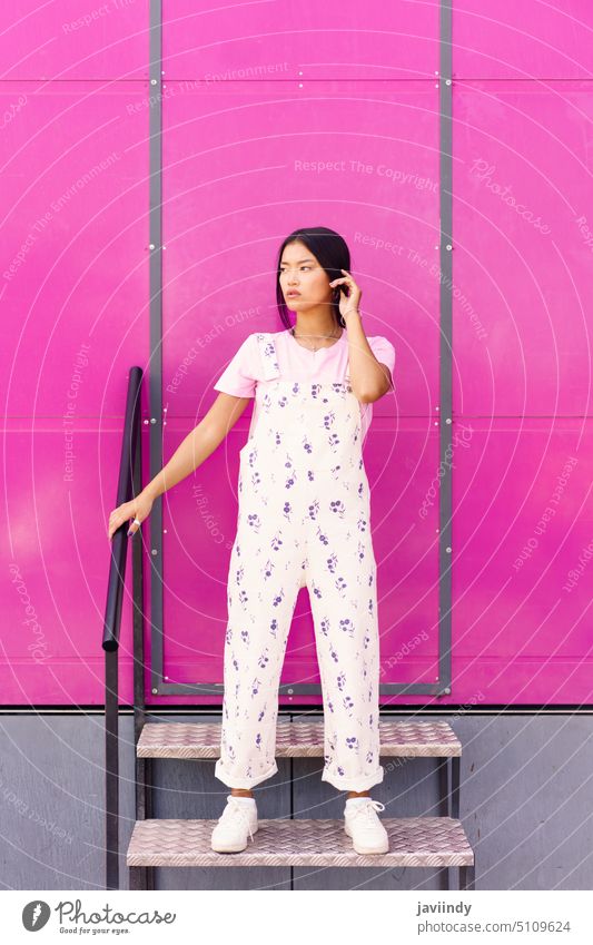 Chinese woman with blank stare and serious expression standing against pink wall of modern building. asian chinese fashionable urban japanese person girl female