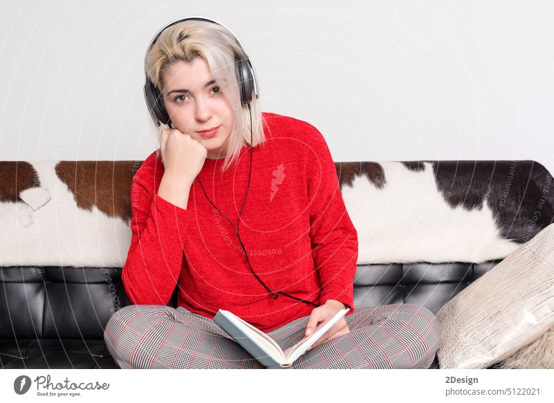 Bored woman with headphones sitting on leather couch holding book blonde reading bored barefoot sweater indoor young sofa home female person caucasian leisure