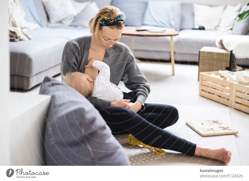 Young woman breastfeeding her infant baby boy casualy sitting on child's playing mat on living room floor at home. parent mother motherhood family milk kid care