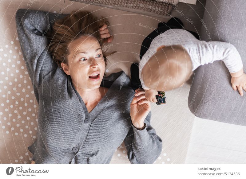 Happy family moments. Mother lying comfortably on children's mat playing with her baby boy watching and suppervising his first steps. Positive human emotions, feelings, joy.