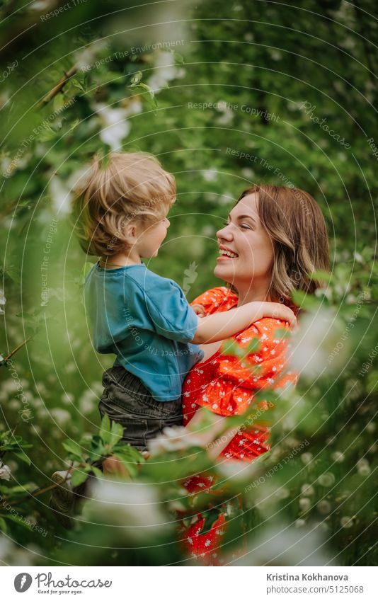 Portrait of happy mother and her baby boy. Beautiful touching scene of mom and son admires each other and sincerely smiles. Blooming garden in spring. Family, kiss, childhood concept.