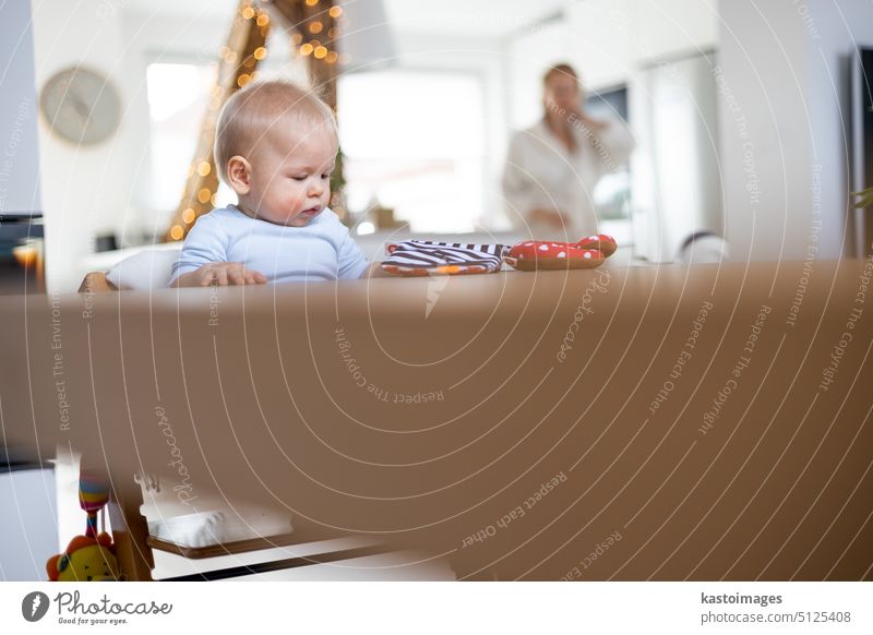 Happy infant sitting at dining table and playing with his toy in traditional scandinavian designer wooden high chair in modern bright atic home superwised by his mother.