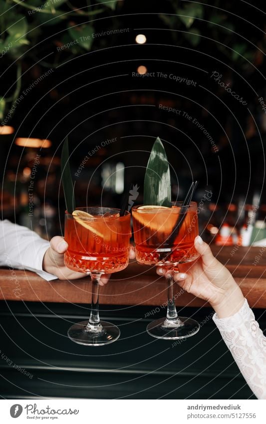 hands of woman and man are clinking, cheers with glasses of Spritz cocktail. Couple celebrating wedding, anniversary with Aperol spritz cocktails, with orange and greens Refreshing alcoholic drink