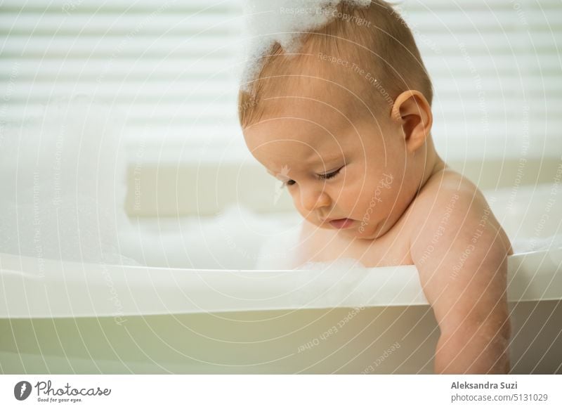 Cute little baby sitting in white bathtub with foam and soap bubbles. Taking bath and playing with toys. Baby hygiene. 6 month 6 month baby adorable bathe