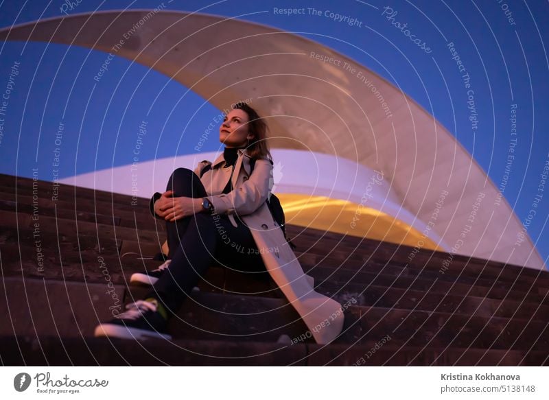 Pretty traveler woman with backpack sitting on stairs of Auditorium in Santa Cruz de Tenerife. Beautiful modern architecture. Portrait of girl relaxing outdoors in city.