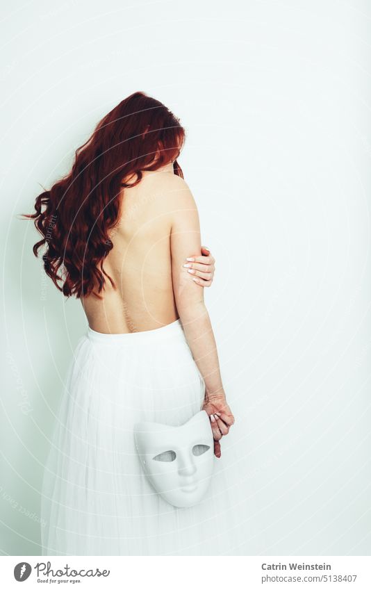 A woman with long red hair stands with her back to the camera. She is topless, wears a white tulle skirt and has a white mask in her hand. Woman Feminine Naked