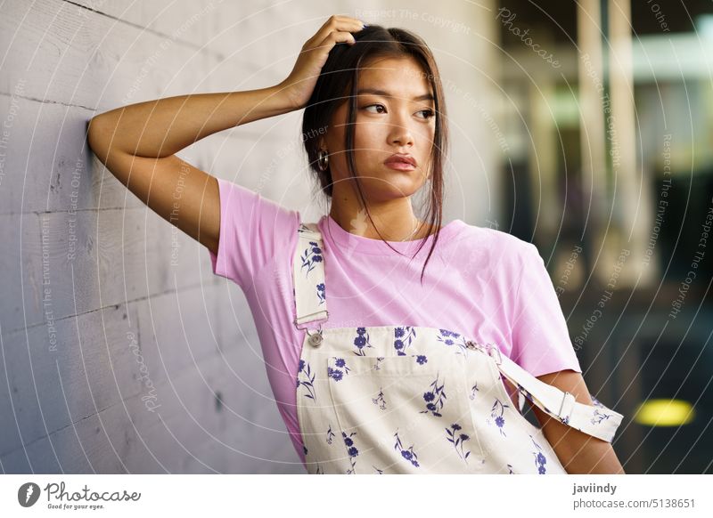 Stylish Asian female leaning on wall woman calm street style urban modern touch head personality young asian ethnic t shirt overall peaceful feminine daytime