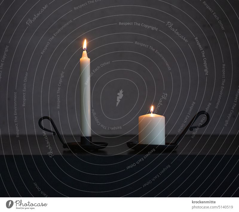 Two candles in black candlesticks with handle burn in front of a concrete wall. Candlelight shoulder stand Thin Fat Wax Candle holder Concrete wall Grief