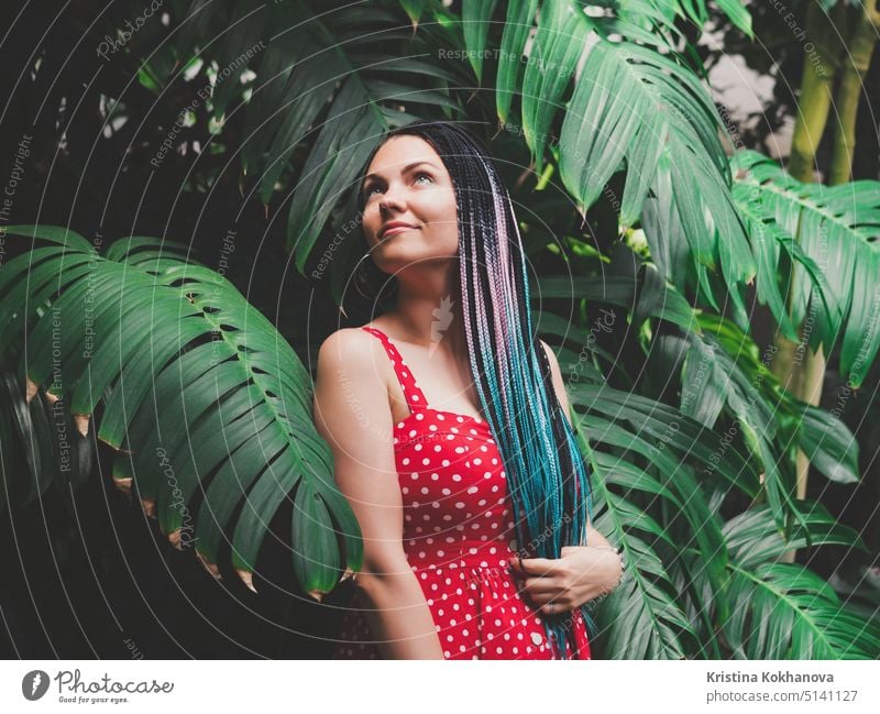 Young pretty girl with unusual hairstyle - colorful long african braids standing in the jungle on tropical leaves background. beautiful green nature people