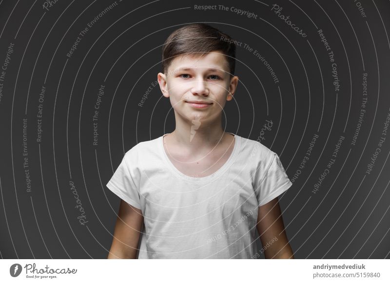 Cute young boy in white T-shirt posing in front of gray empty wall. Portrait of fashionable male child. Smiling boy posing, blank wall on background. Concept of children style and fashion