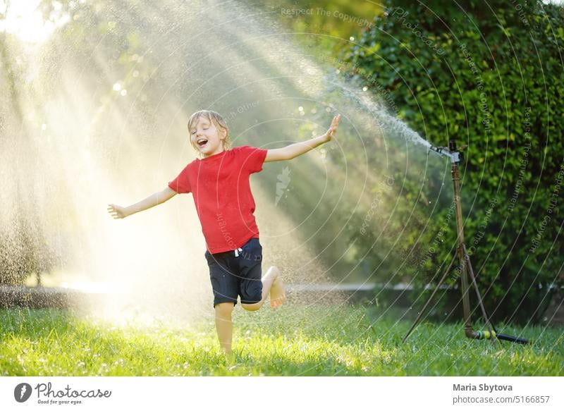 Funny little boy playing with garden sprinkler in sunny backyard. Elementary school child laughing, jumping and having fun with spray of water. Summer outdoors activity for kids.