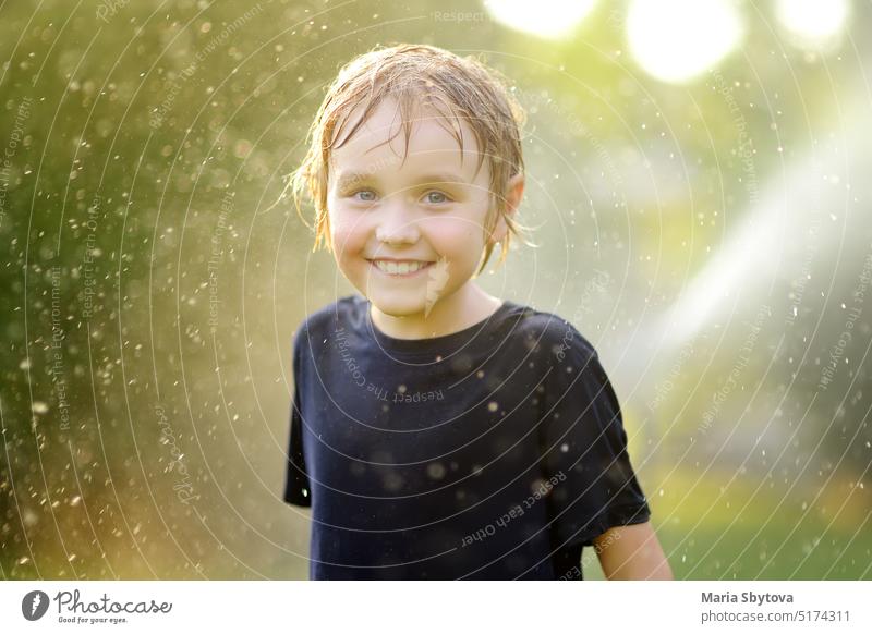 Close-up portrait of funny little boy playing with garden sprinkler in sunny backyard. Elementary school child laughing, jumping and having fun with water. Summer outdoors activity for kids.