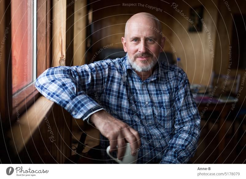 a man with a beard and a check shirt sits looking contented in a simple wooden house at the window with a coffee cup in his hand. Man Facial hair kind