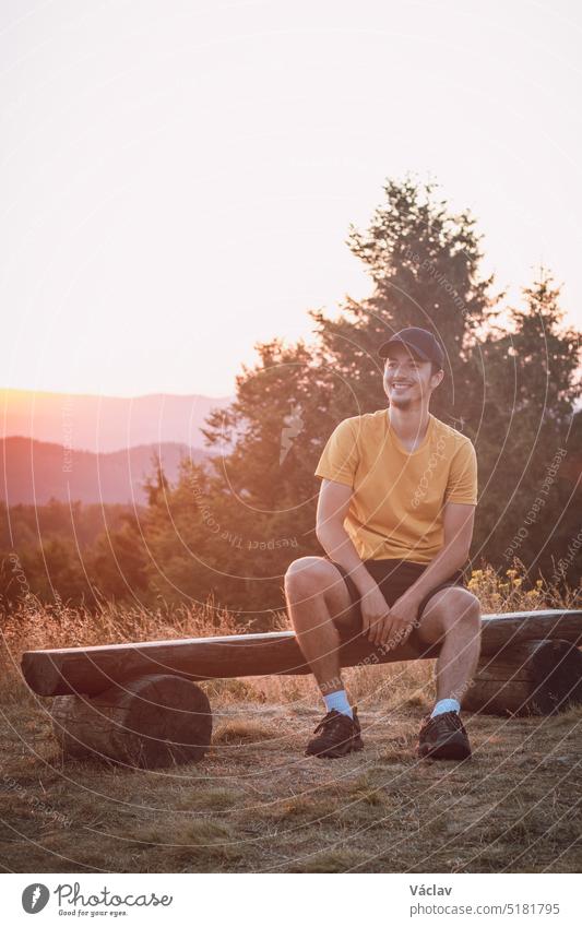 Enthusiastic traveler smiles and sits on a wooden bench, watching the last remnants of sunshine in the raw nature. Sunset on Horni Becva, Beskydy mountains, Czech republic