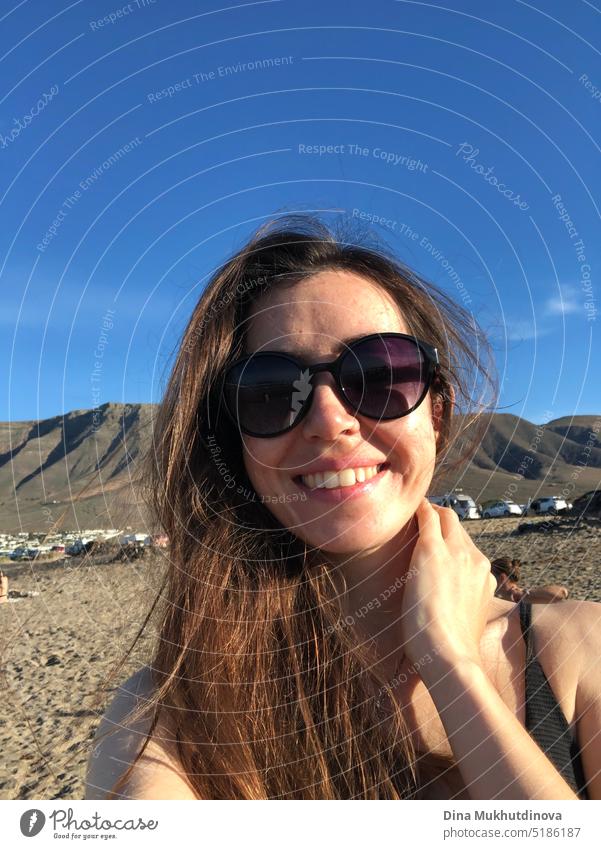 Selfie of a girl wearing sunglasses. Young woman relaxed on vacation, standing on the beach near mountains, in sunset light, smiling. Real people. Authentic image of real people.