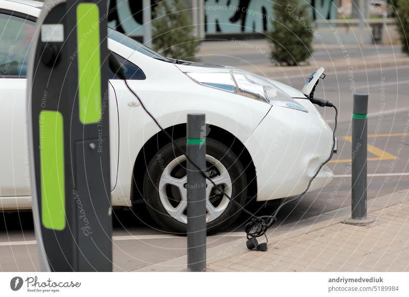 Charging modern electric cars, new energy vehicles, NEV, on the street white electric car with a cable connected and a charging station. Eco-friendly alternative energy concept.