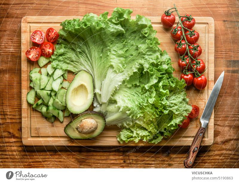 Healthy fresh salad ingredients: lettuce, halved avocado, cucumber and tomatoes on wooden cutting board with knife, top view healthy lunch cooking raw
