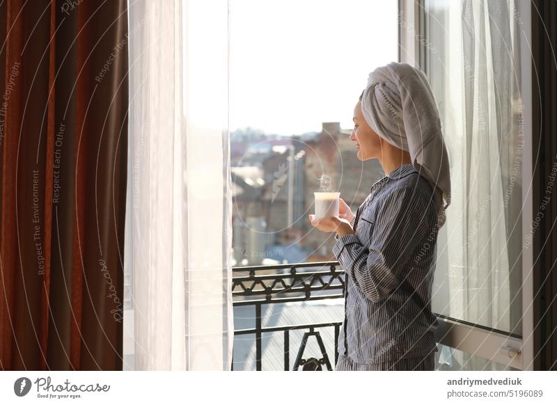 smiling young woman in nightwear and towel on head is holding cup of hot coffee or tea on balcony by the window in the morning. Concept of domestic lifestyle.