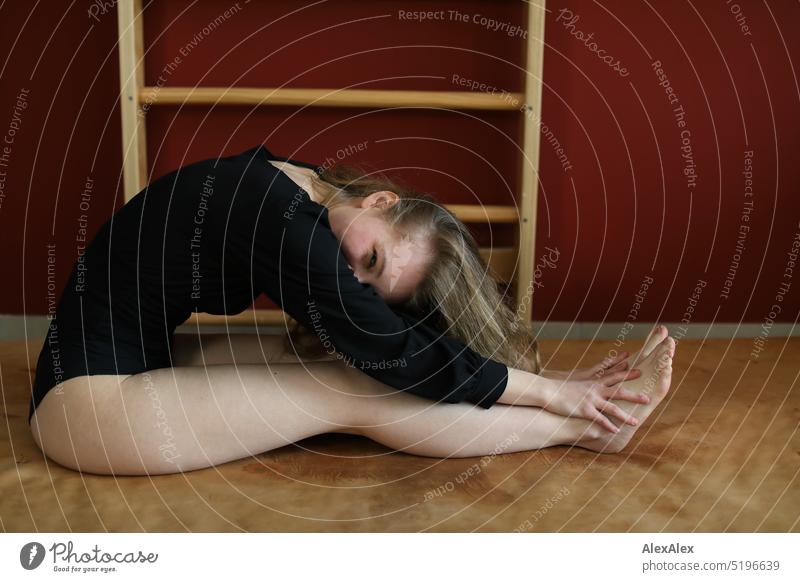 a young blonde slim woman in black bodysuit is doing gymnastics on a gym mat in front of a red wall with wall bars Woman youthful Slim Athletic pretty Smiling