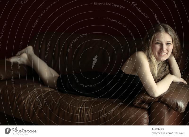 young blonde slim woman in black dress lies barefoot on brown leather couch and smiles Woman youthful Slim Athletic pretty Smiling fit Adults Esthetic naturally