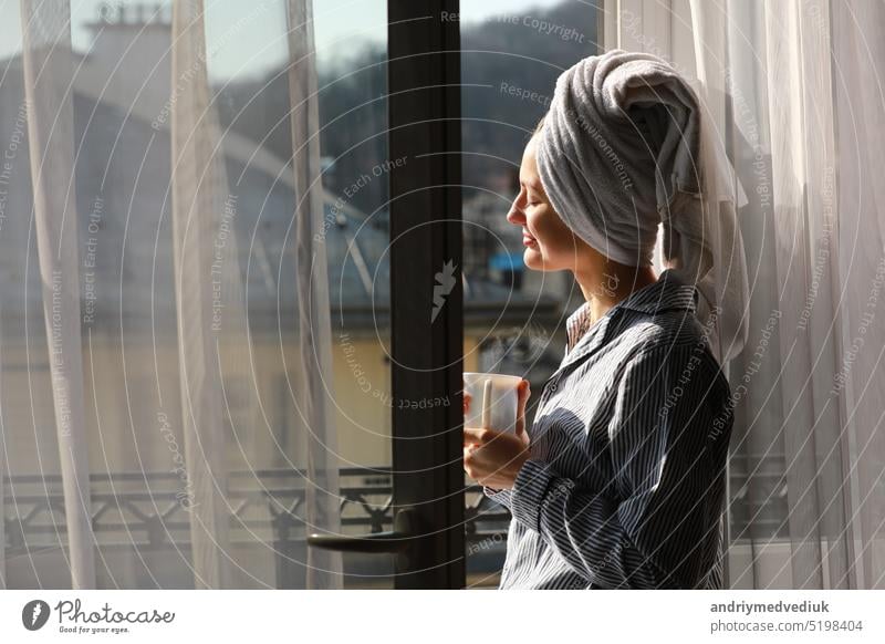 Beautiful young woman with bath towel on head holds a white cup of hot coffee or tea at home by the window. Smiling girl after shower is enjoying aroma drinks and have breakfast in the morning