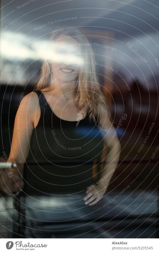 Portrait of blonde long haired woman in black tank top behind window pane Woman portrait Blonde Long-haired Smiling Looking into the camera Slim daintily pretty