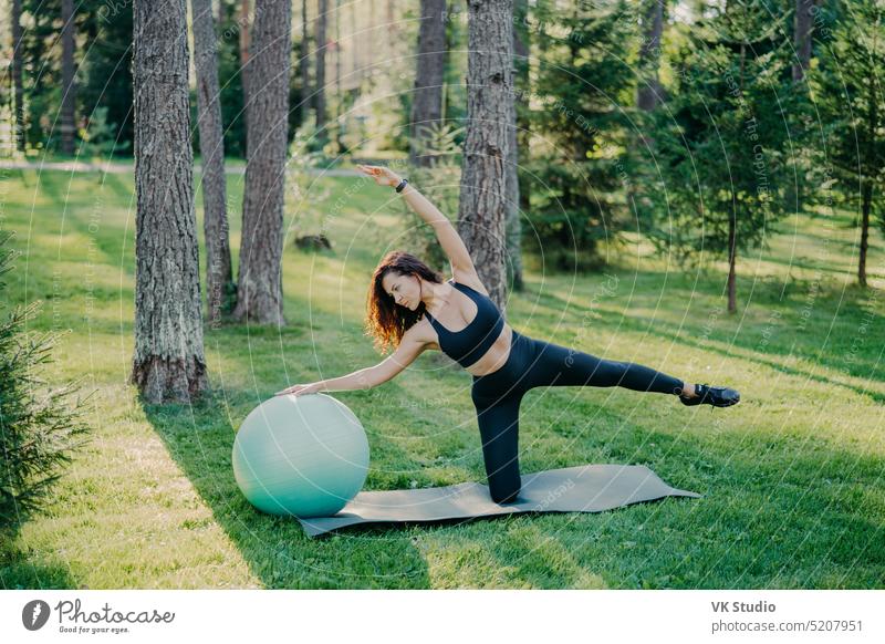 Sport and active lifestyle concept. Slim sporty woman in black clothing leans aside on fitness ball, raises arms, poses on karemat in green park, trains yoga outdoor, has athletic body shape.