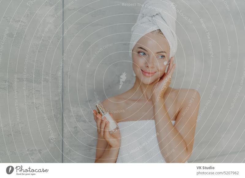 Lovely young woman applies beauty lotion on face has satisfied expression wears bath towel on head poses with bare shoulders isolated over grey background. Cosmetology wellbeing and spa concept