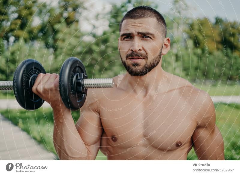 Serious sportsman poses with naked torso has muscular body raises dumbbell, poses outdoor, being in good physical shape, enjoys fresh air and nature. Strength, sporty lifestyle and bodybuilding