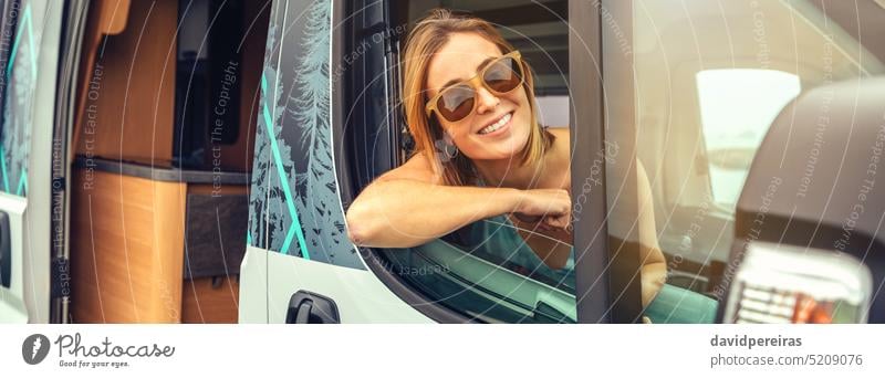 Young woman in sunglasses sitting in camper van leaning on the door happy young banner web header panorama panoramic copy space summer trip travel smiling