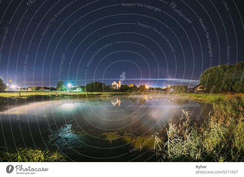 Night Clouds Over River Lake Near Houses In Village. Night Starry Sky Above Lake River With Bright Stars And Meteoric Track Trail. Glowing Stars Above Summer Nature