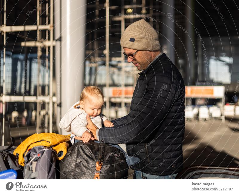 Fatherat comforting his tired infant baby boy child sitting on top of luggage cart in front of airport terminal station while traveling wih family. father dad