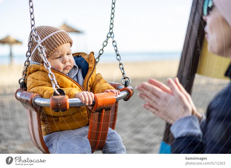 Mother pushing her infant baby boy child on a swing on sandy beach playground outdoors on nice sunny cold winter day in Malaga, Spain. happy son family kid