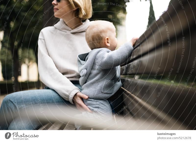 Young mother with her cute infant baby boy child sitting on bench in city park son young lifestyle love little family woman together cheerful play stand happy