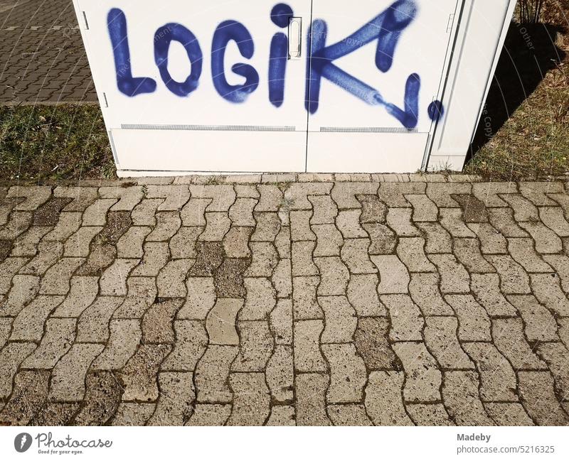 The word LOGIK in blue writing as graffiti on a bright distribution box on the sidewalk with interlocking sidewalk in sunshine in Oerlinghausen near Bielefeld on the Hermannsweg in the Teutoburg Forest in East Westphalia Lippe