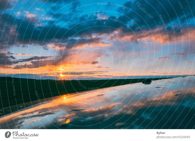 Sunset Sky With Setting Sun Reflected In Hood Of Car. Countryside Landscape Under Scenic Colorful Sky At Sunset Dawn Sunrise. Skyline amazing background