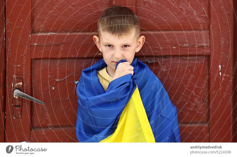 Ukrainian small child stands outdoors supporting homeland, little kid covered in national flag looking at camera. Protesting against Russian war in Ukraine, armed conflict, homeless, crisis, portrait