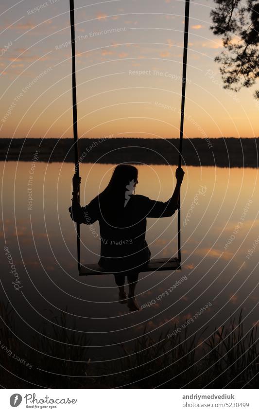 silhouette of a romantic young woman on a swing over lake at sunset. Young girl traveler sitting on the swing in beautiful nature, view on the lake outdoor sky