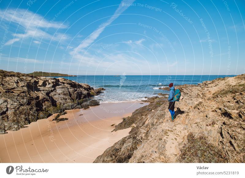 Backpacker walks along a rock and across a sandy beach on the marked Fisherman Trail on the Atlantic coast near Porto Covo, Portugal. Rota Vicentina student