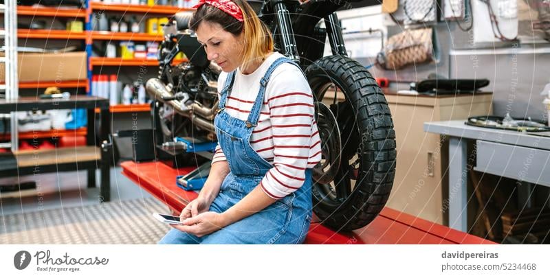Mechanic woman looking phone sitting over platform with motorcycle on factory mechanic female using cellphone telephone smiling happiness motorbike portrait