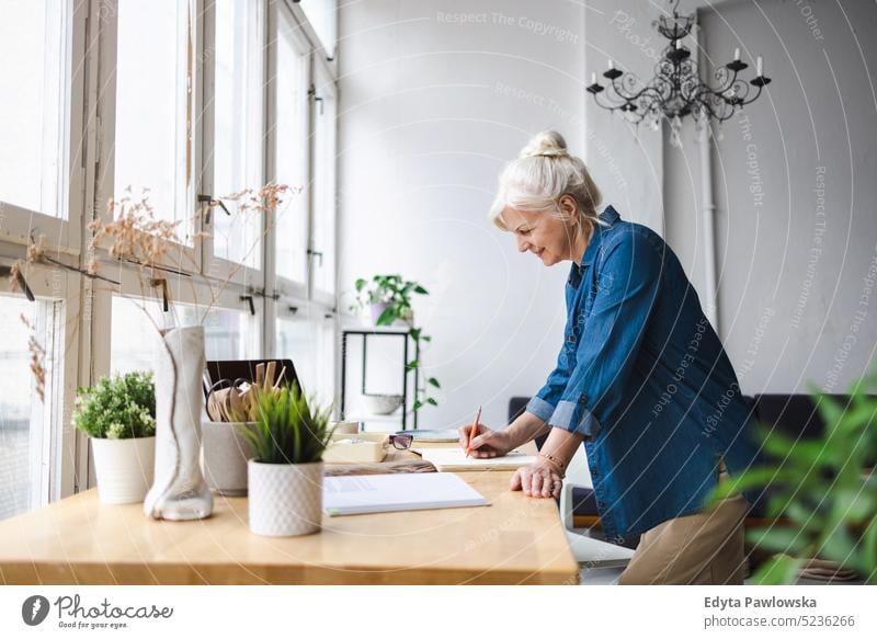 Smiling mature businesswoman writing in notebook while sitting at table in office real people senior indoors loft window home mature adult one person attractive