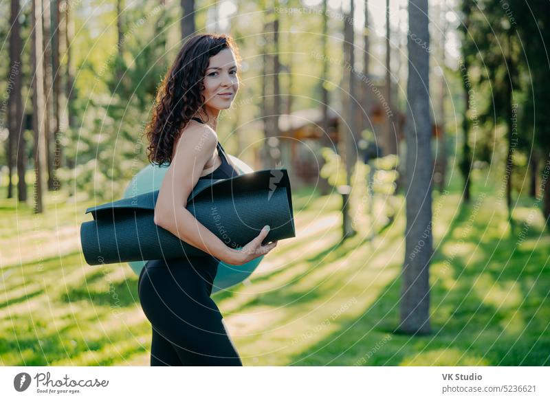 Sideways shot of sporty slim young woman dressed in black leggings, carries rolled up karemat under arm, poses against green grass and trees background, going to have exercises with fitness ball