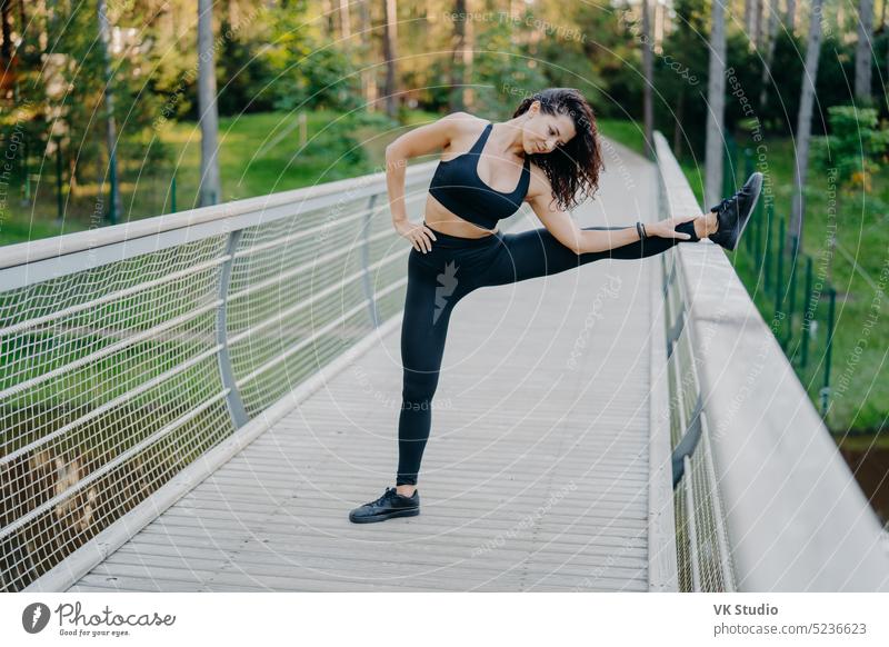 Determined sportswoman stretches legs on bridge, wears active wear, demonstrates good flexibility, poses outdoor against forest background. Woman runner prepares for jogging, has summer workout