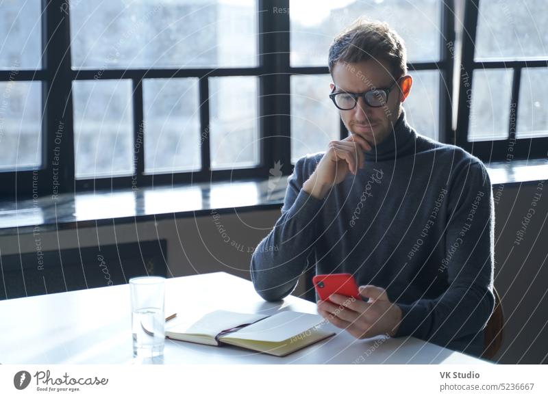 Young german businessman reading internet news or checking email on smartphone while sitting at desk young mobile table office sms hold call austrian notebook