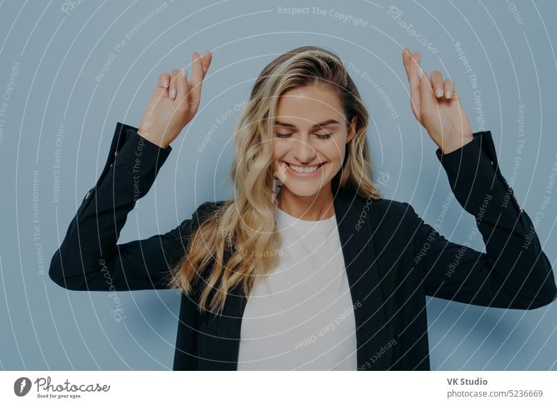 Young blonde woman making wish with crossed fingers young excited black coat holding both hands air wishing good luck crossing hope expression smile face